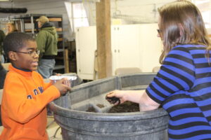 A woman in a blue striped shirt with soil in her hands and a child in an orange shirt holding a small metal tray stand on opposite sides of a large tub of soil