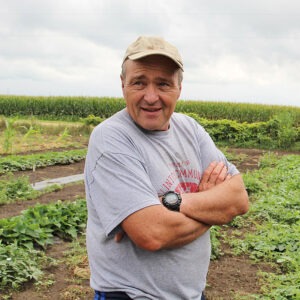 man wearing cap and T-shirt and crossing arms stands in the middle of a field of crops