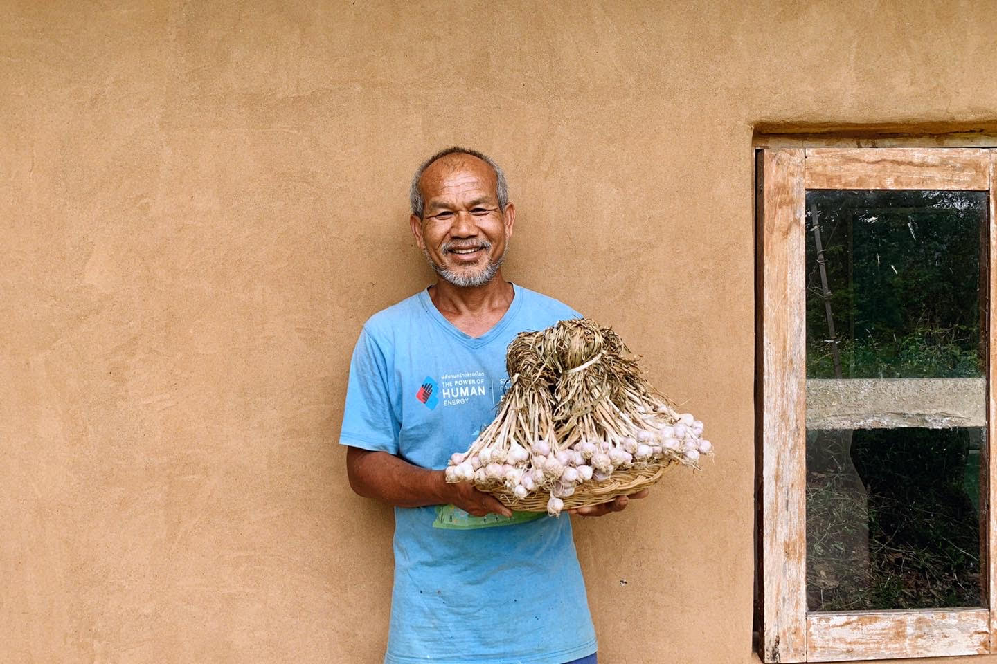 A man holding a basket full of garlic bulbs standing against a tan wall.
