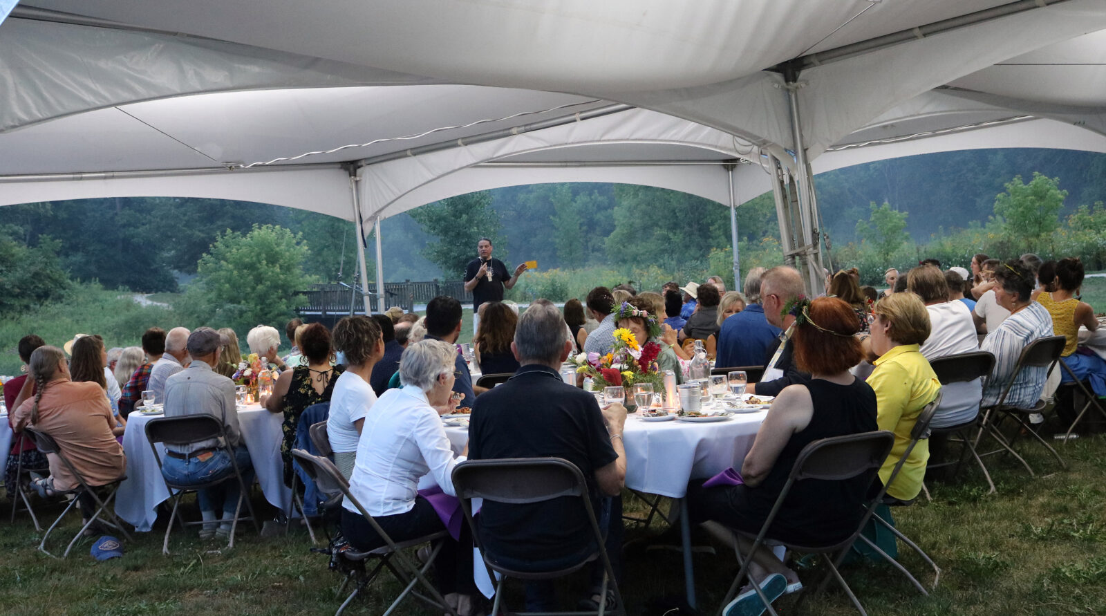 A large group of people sit at tables under a white tent at a dinner